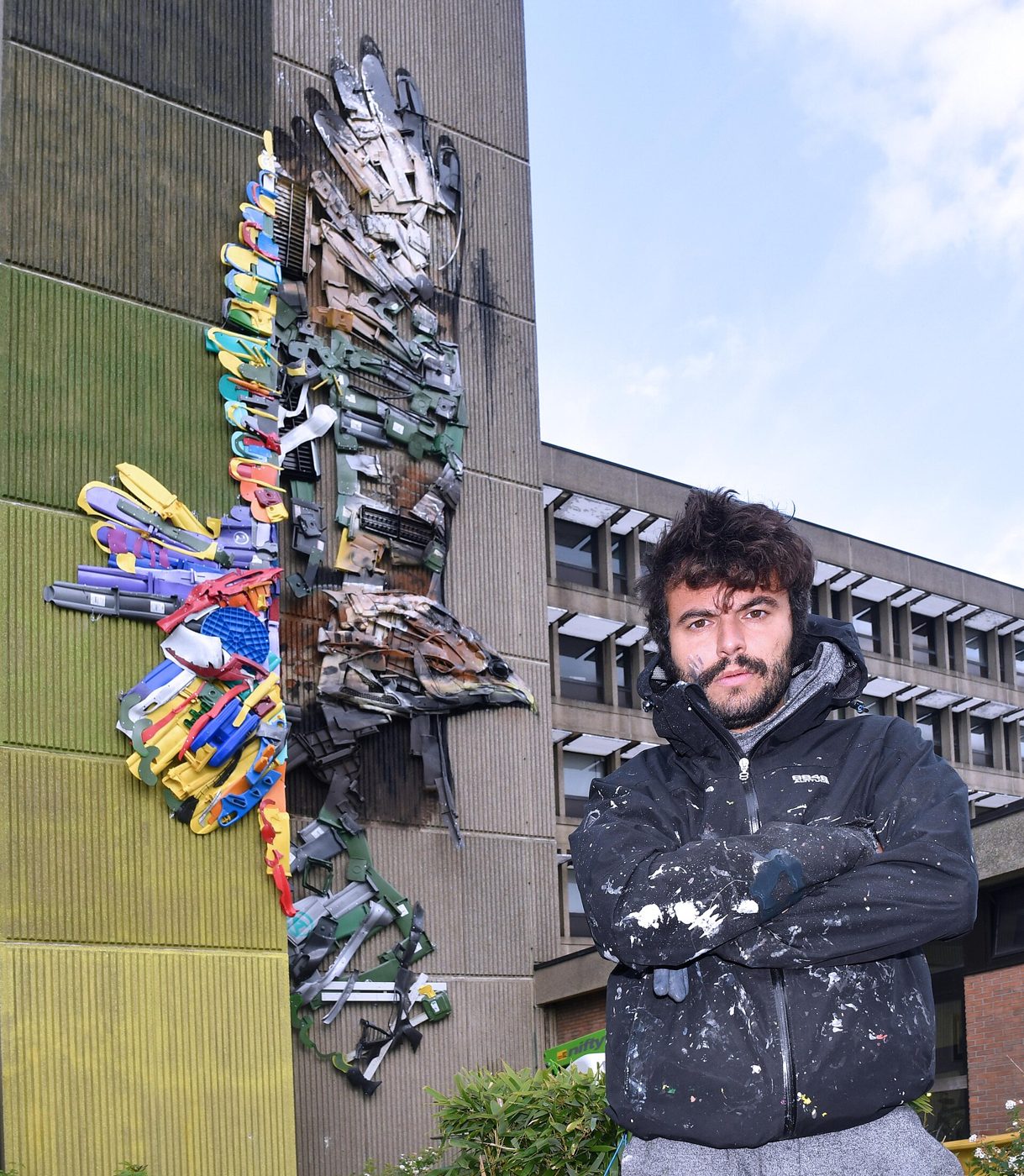 Bordalo II, de casaco escuro, ao lado de um relevo colorido de resíduos numa fachada