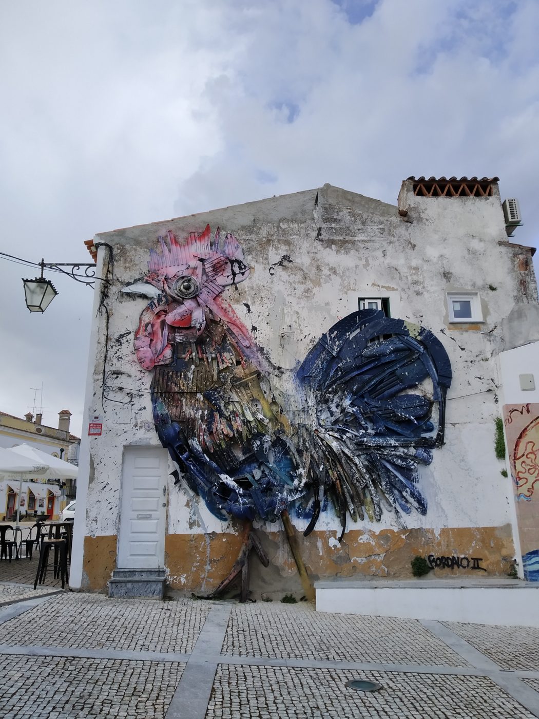 Galo enorme em relevo numa parede branca, com penas de plástico e metal coloridas