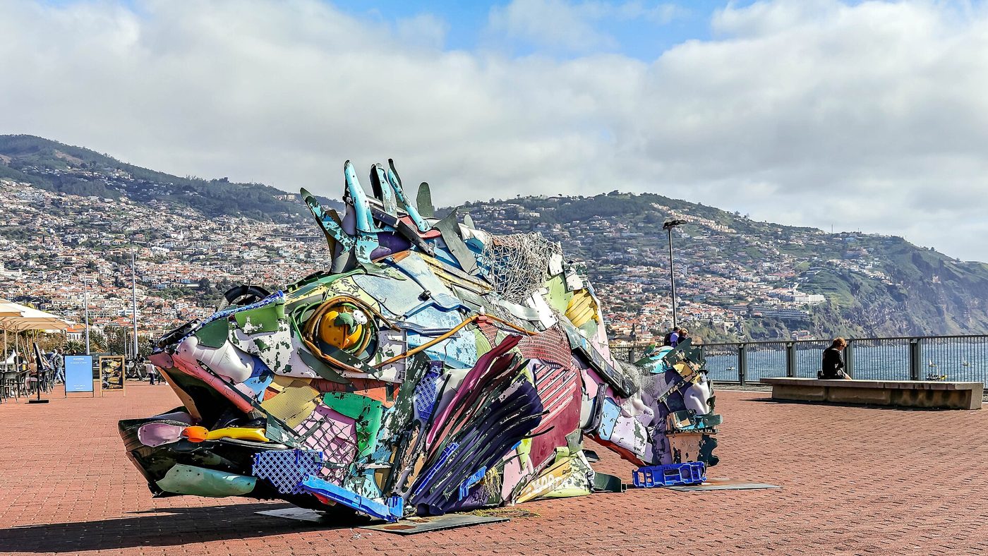 Peixe enorme feito de plásticos coloridos numa praça junto ao mar, com a cidade do Funchal ao fundo
