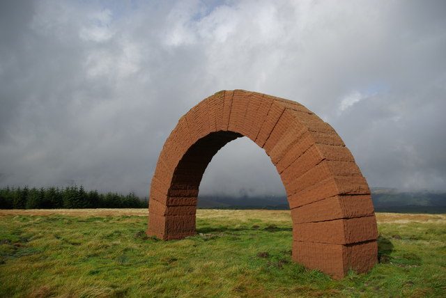 Arco de pedra vermelha numa colina coberta de erva, sob céu nublado