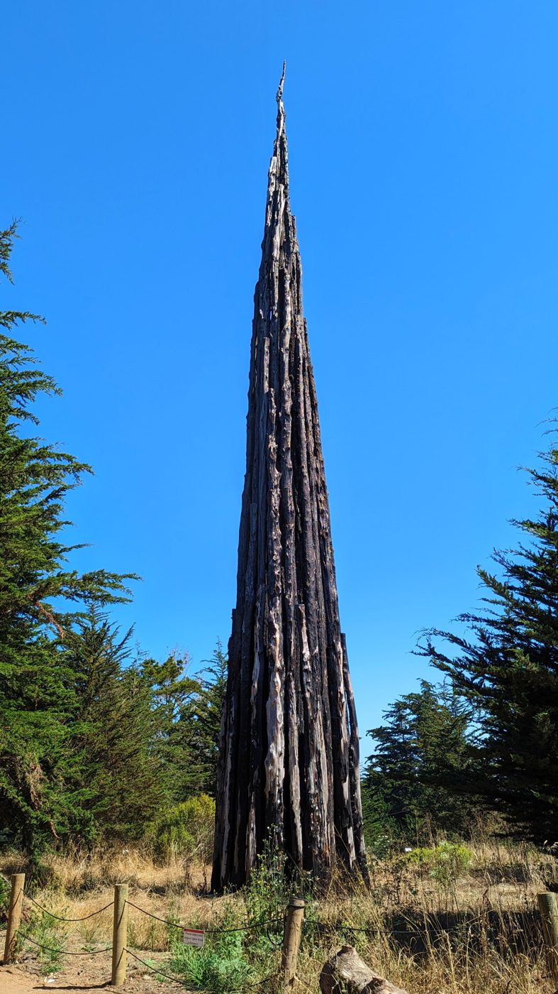 Escultura alta e pontiaguda feita de troncos de árvore, a erguer-se contra o céu azul