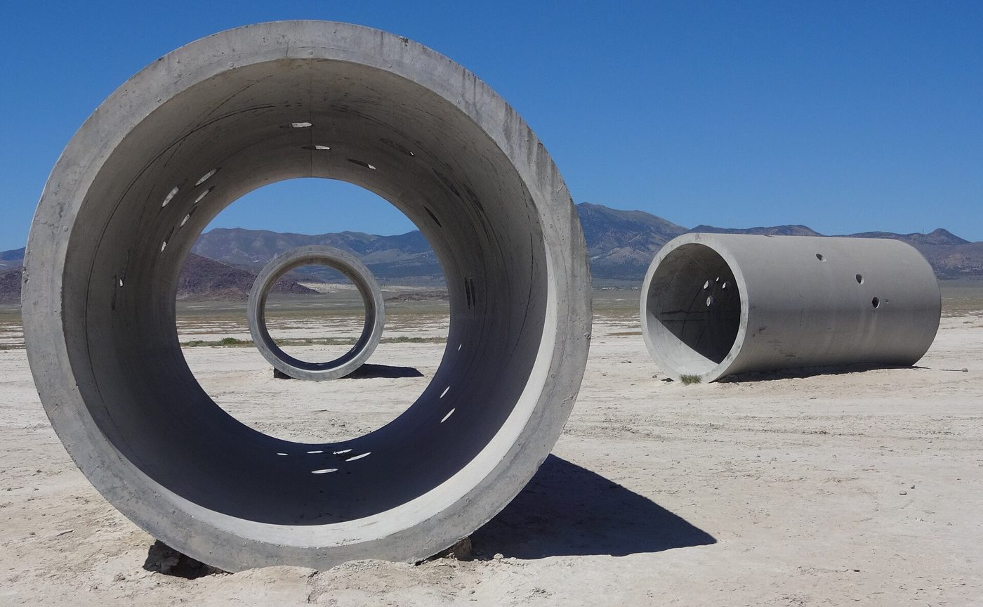 Tubos de betão gigantes pousados no deserto; através de um deles vê-se outro tubo e as montanhas