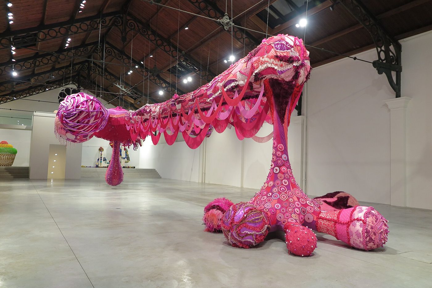 Grande escultura têxtil cor-de-rosa, alongada, pousada no chão de um pavilhão