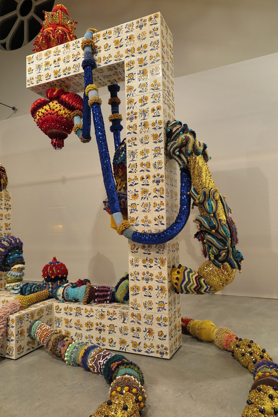 Escultura com azulejos, objetos suspensos e longas formas cobertas de croché colorido